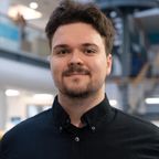 Head shot of Imre Draskovits, smiling, with short brown hair and a beard, wearing a black shirt, in a bright university atrium.