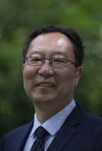 Head shot of Li Wei, smiling, wearing glasses, a dark suit and tie, in front of green foliage.