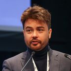 Michael Barngrover speaking at a conference microphone, with short brown hair and a beard, wearing a dark jacket and roll-neck.